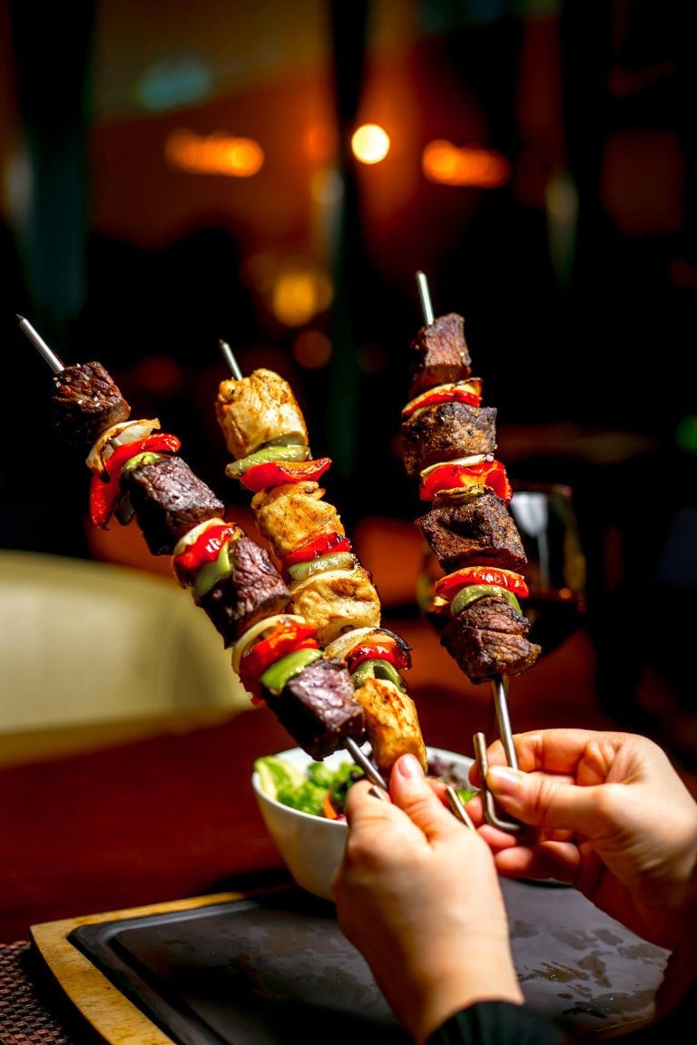 side view of hands holding shish kebab with colorful bell peppers and fresh vegetable salad on a wooden table